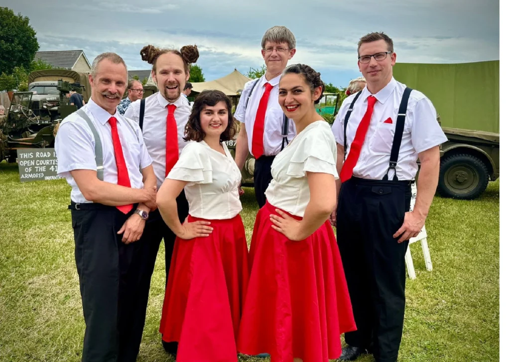 Six performers from the East Anglian Swing Troupe pose outdoors at a 1940s-themed event. The three men wear white shirts, red ties, black trousers, and braces. The three women wear white blouses with red flared skirts and red lipstick. Behind them are vintage military vehicles, including a jeep, and a green tent. A sign nearby reads “THIS ROAD IS OPEN COURTESY OF THE 41st ARMORED INFANTRY.”