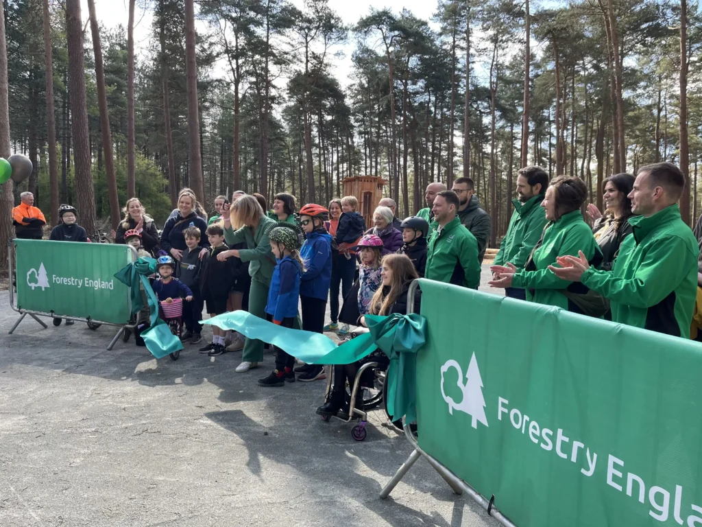 A large group of adults and children—some in helmets and cycling gear—gather outdoors among tall pine trees for a ribbon-cutting event hosted by Forestry England. Green Forestry England barriers line both sides, and a wide green ribbon is being cut by a woman in the centre. Smiling onlookers and forest rangers in green jackets applaud.