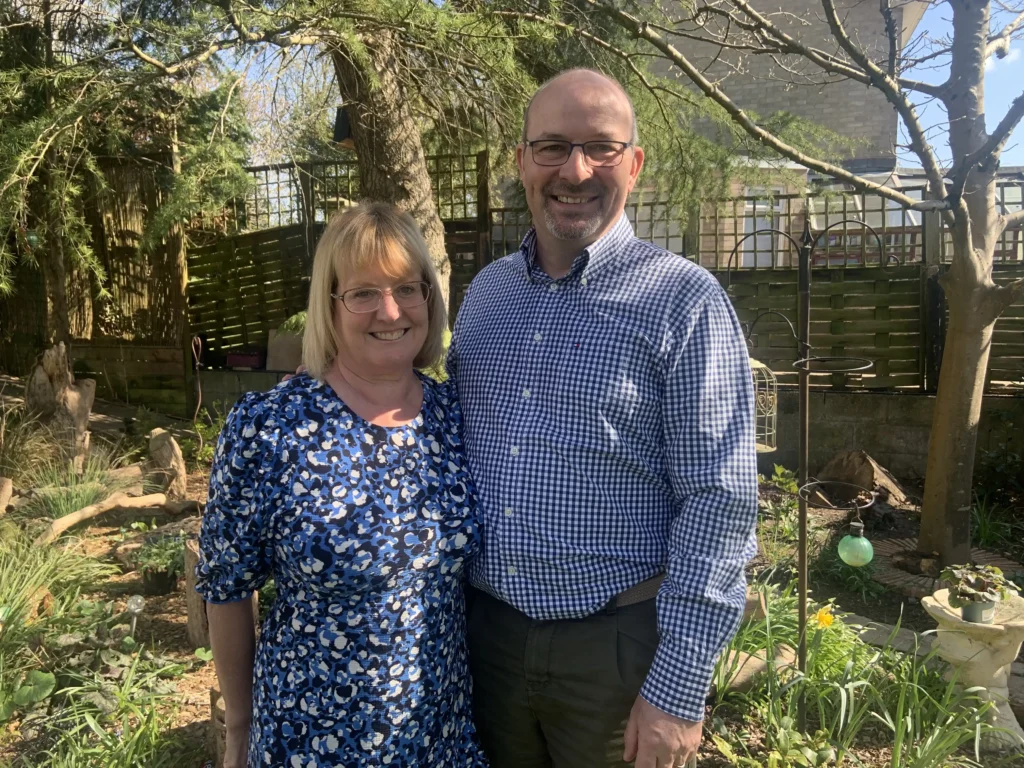 A smiling man and woman (identified as Peter and Corrina Turner) stand together in a garden with greenery and flowers around them. The woman wears a blue floral dress and glasses, and the man wears a blue checked shirt and glasses. It’s a sunny day with trees casting light shadows.