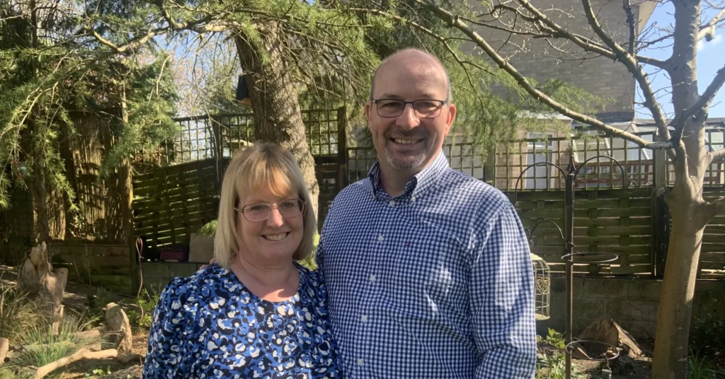 A smiling man and woman (identified as Peter and Corrina Turner) stand together in a garden with greenery and flowers around them. The woman wears a blue floral dress and glasses, and the man wears a blue checked shirt and glasses. It’s a sunny day with trees casting light shadows.