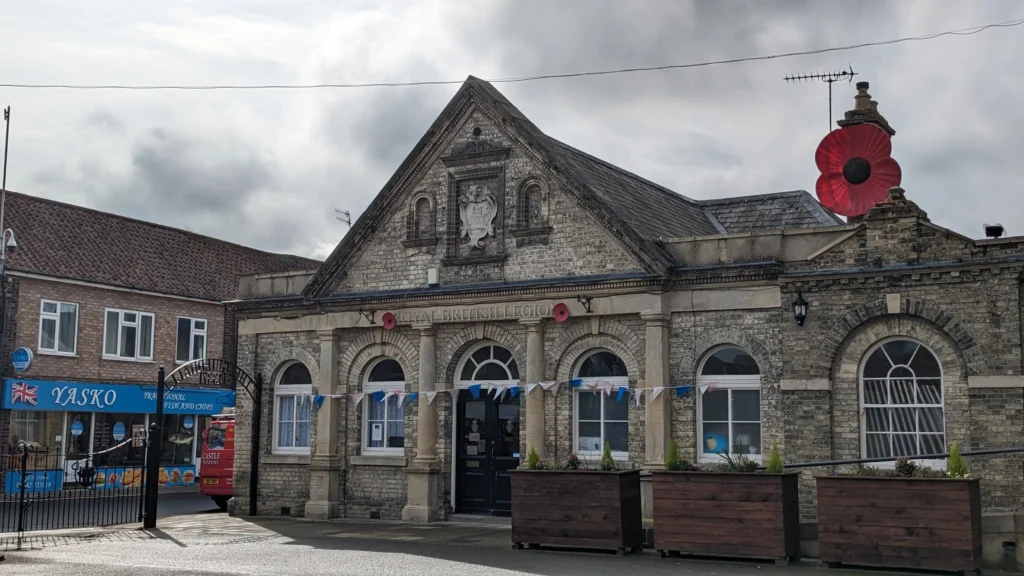 The Royal British Legion building in Thetford, a historic brick structure with arched windows and poppy decorations on the facade. Blue and white bunting hangs across the front, and large planters line the pavement. The sky is overcast, giving a moody atmosphere.