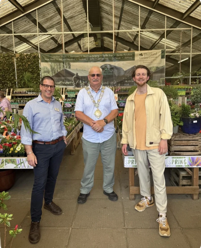Three men stand smiling inside Thetford Garden Centre, surrounded by colourful spring plants and flowers. In the centre is the Mayor of Thetford, Chris Harvey, wearing a light blue shirt and his ceremonial mayoral chain. On the left is Julian Chittock in a blue shirt and dark trousers, and on the right is Joey La Meche in a cream jacket and trainers. Behind them, a banner reads “Thetford Garden Centre” and lists various gardening departments.
