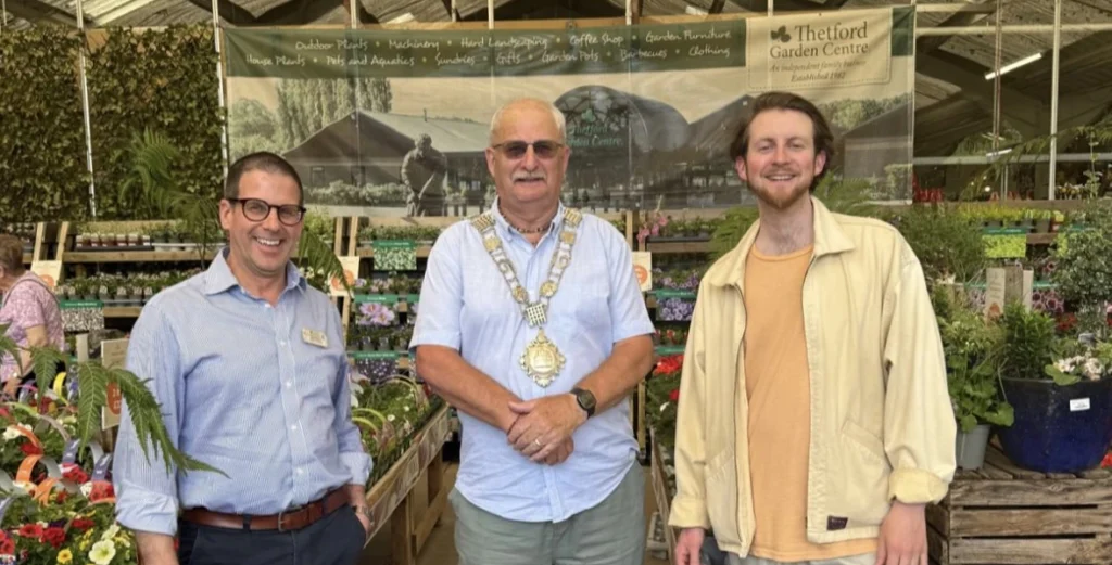 Three men stand smiling inside Thetford Garden Centre, surrounded by colourful spring plants and flowers. In the centre is the Mayor of Thetford, Chris Harvey, wearing a light blue shirt and his ceremonial mayoral chain. On the left is Julian Chittock in a blue shirt and dark trousers, and on the right is Joey La Meche in a cream jacket and trainers. Behind them, a banner reads “Thetford Garden Centre” and lists various gardening departments.