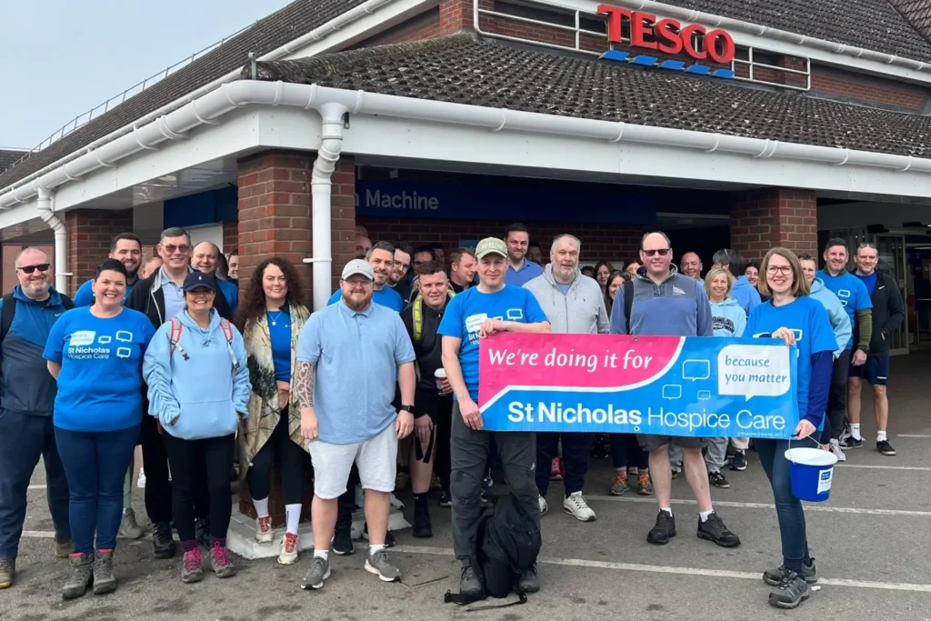 A large group of people gathered outside a Tesco store, many wearing blue St Nicholas Hospice Care T-shirts and hoodies. The group is posing together in the car park before a fundraising walk. At the front, several participants are holding a bright banner that reads: "We're doing it for St Nicholas Hospice Care" with the charity’s “because you matter” slogan. One person holds a donation bucket. The atmosphere is upbeat and community-focused.