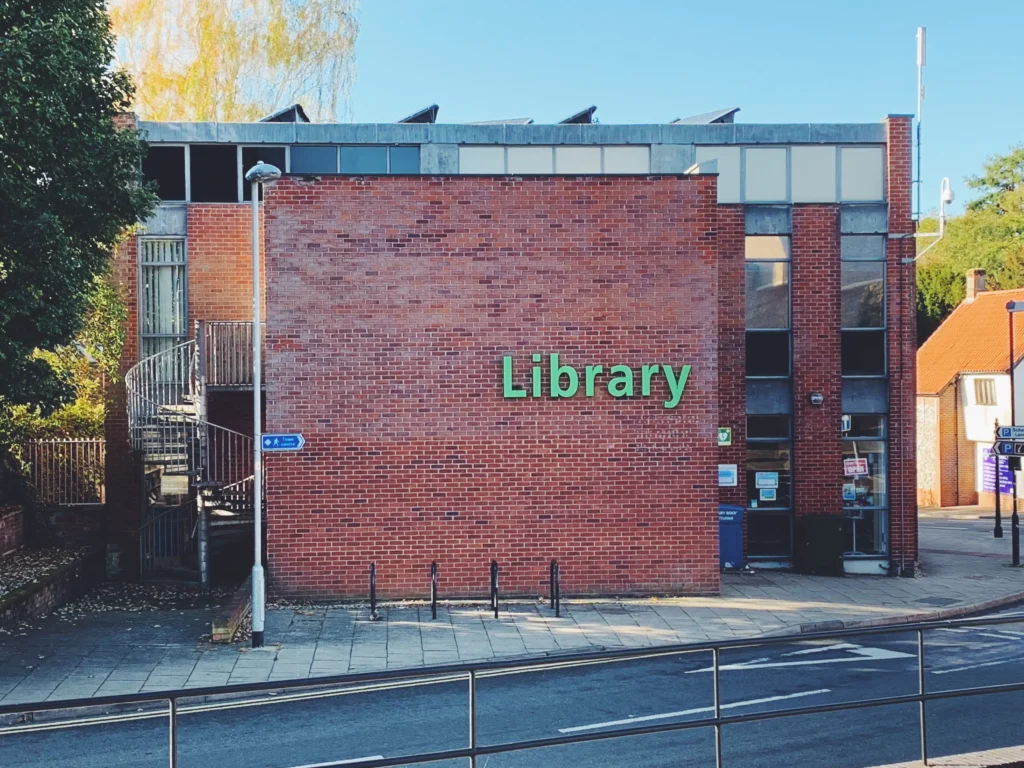 The exterior of Thetford Library, a modern red-brick building with large windows and a green "Library" sign on the central wall. A metal spiral staircase is visible on the left side, and there's a streetlamp and pavement in front. A signpost directs pedestrians to nearby points of interest. The sky is clear and blue, and autumn leaves are scattered near the base of the building.