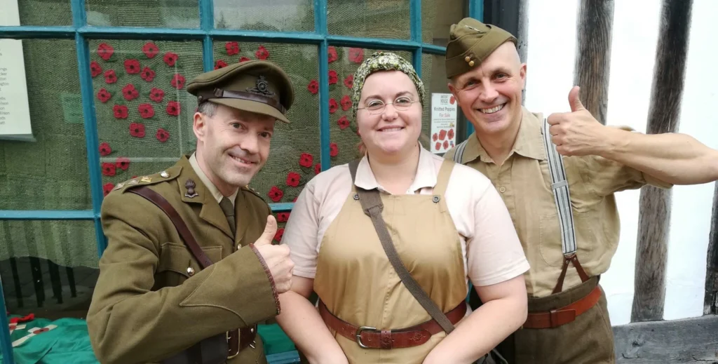 Three people in WWII-era costumes smile and give thumbs up in front of a timber-framed building. The two men wear military uniforms, while the woman in the middle wears beige overalls and a floral headscarf. The window behind them is decorated with red poppies.