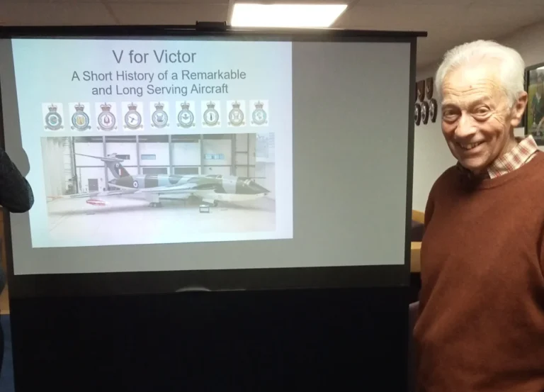 An older man stands smiling beside a projector screen displaying a presentation titled “V for Victor: A Short History of a Remarkable and Long Serving Aircraft.” The slide shows a photo of a Victor bomber and the badges of various RAF squadrons. The man wears a rust-coloured jumper and stands in a room with blue carpet, wooden tables, and ceiling lights. A laptop and projector are visible in the foreground.
