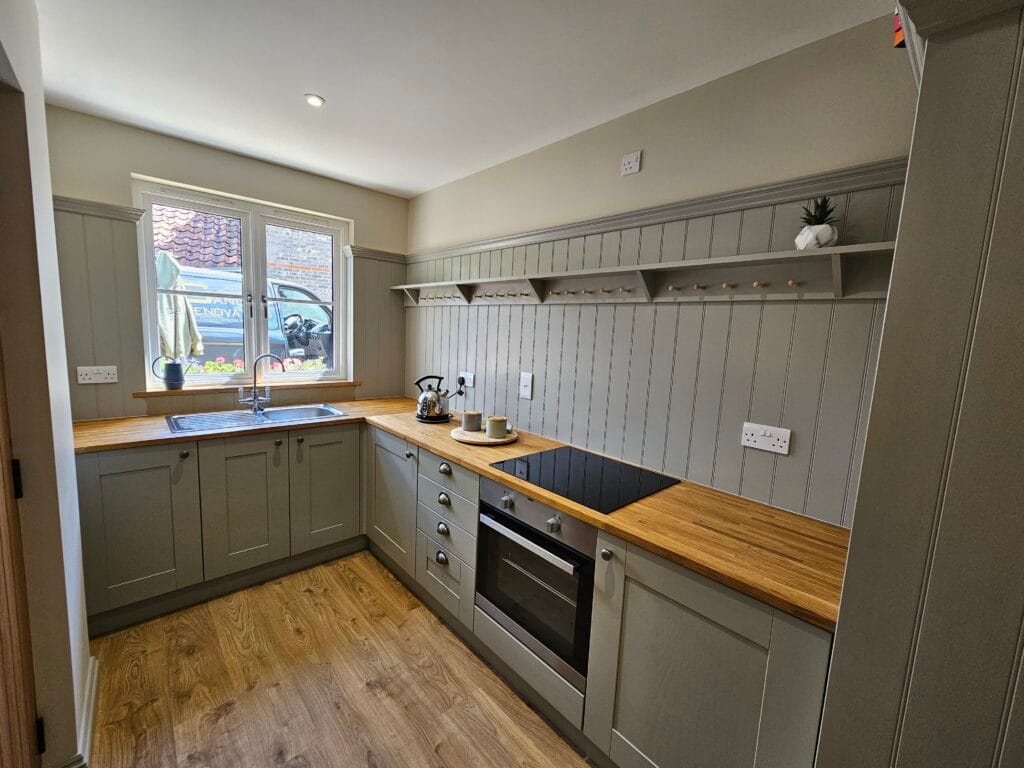 A professionally renovated kitchen featuring wooden countertops, grey shaker cabinets, a built-in oven, and a wood-effect floor. The walls are half-panelled in grey, with a matching open shelf above. The window brings in natural light, with a kettle and mugs set on the counter.