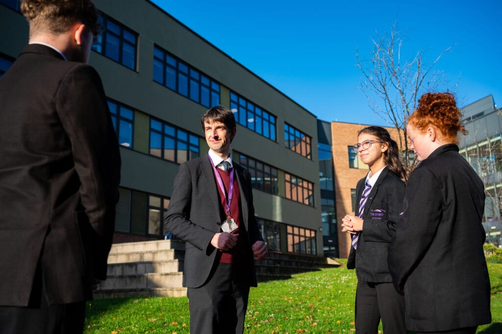 A group of four individuals, including one adult male and three students in Thetford Academy uniforms, stand outside a modern school building on a bright sunny day. The adult, wearing a lanyard and dark suit, appears to be talking to the students, who are listening attentively. The background shows large windows and a neat green lawn with scattered autumn leaves.