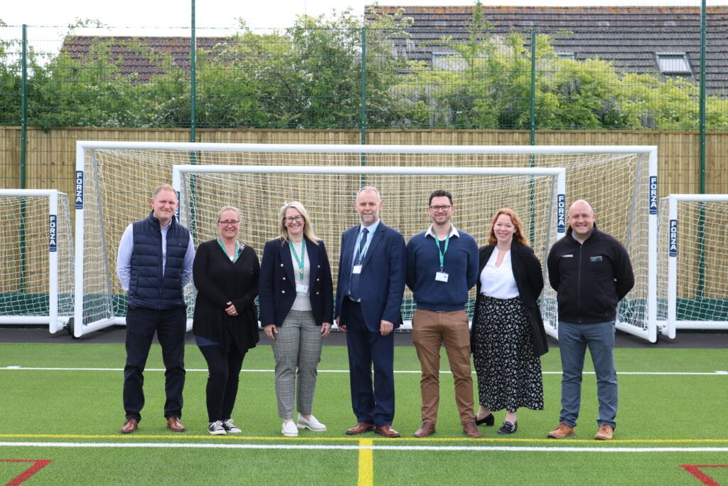 Seven people stand in a row on an outdoor artificial turf sports pitch, with multiple white football goals behind them. The group includes four men and three women, dressed in a mix of business and casual attire, all wearing lanyards. Behind the goals is a tall fence and some greenery, with rooftops visible in the background.