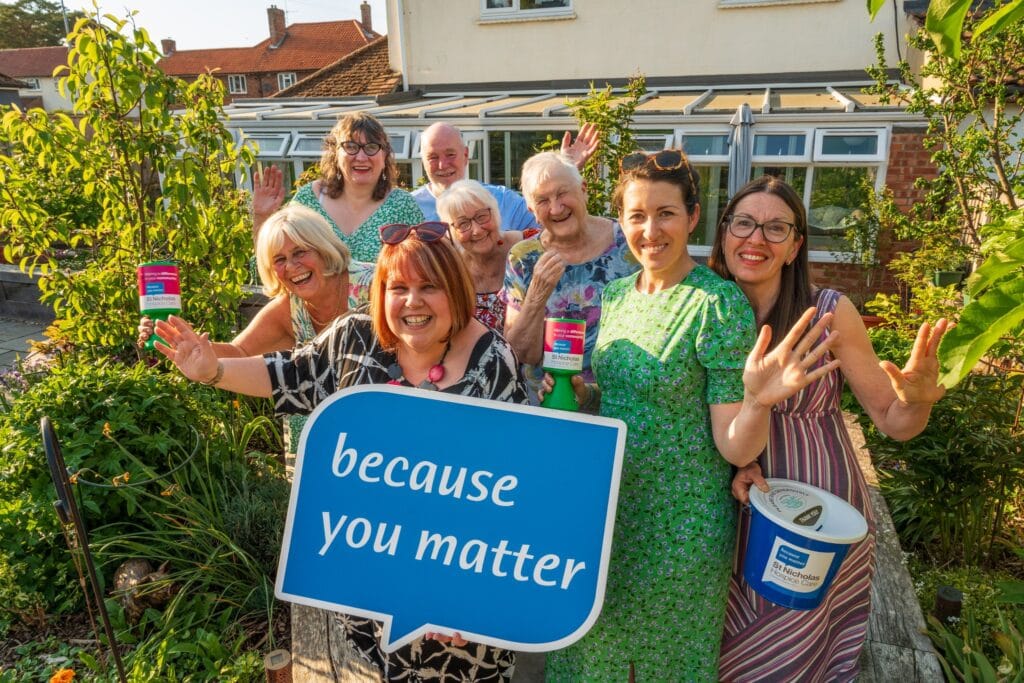 A group of cheerful volunteers stand together in a sunny garden, smiling and waving at the camera. They hold St Nicholas Hospice Care donation buckets and collection tins, with one person at the front holding a large blue sign that reads “because you matter.” The group is surrounded by green plants and flowers, with a conservatory and house visible in the background.