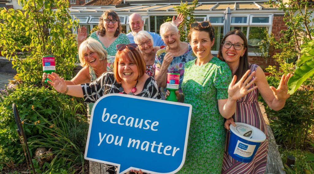 A group of cheerful volunteers stand together in a sunny garden, smiling and waving at the camera. They hold St Nicholas Hospice Care donation buckets and collection tins, with one person at the front holding a large blue sign that reads “because you matter.” The group is surrounded by green plants and flowers, with a conservatory and house visible in the background.