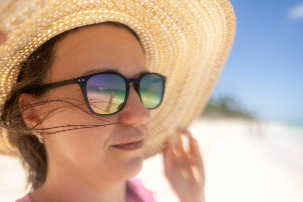  A woman wearing a wide straw sunhat and dark sunglasses stands on a bright sandy beach. Her sunglasses reflect a view of the shoreline and sea. Her face is partially shaded, with loose strands of hair blowing in the breeze under a clear blue sky.