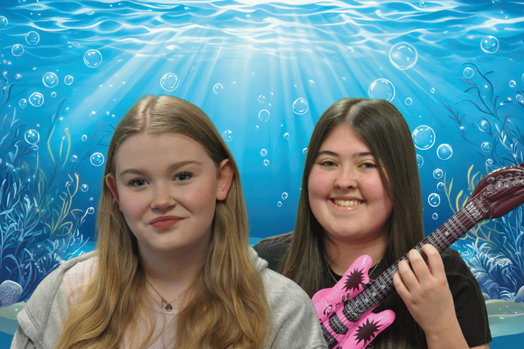 Two teenage girls pose in front of an illustrated underwater backdrop with sunlight filtering through water, seaweed, and bubbles. The girl on the left has long blonde hair and wears a pale hoodie and heart necklace, while the girl on the right has long dark hair and holds a pink inflatable guitar. This promotional photo is for Thetford Players Youth’s upcoming production of The Spongebob Musical: Youth Edition.