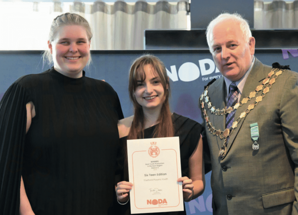 a photo showing two young women, Nadia (left) and Rowan (centre), receiving a certificate from NODA President Graham Liverton (right) in front of a blue NODA backdrop.