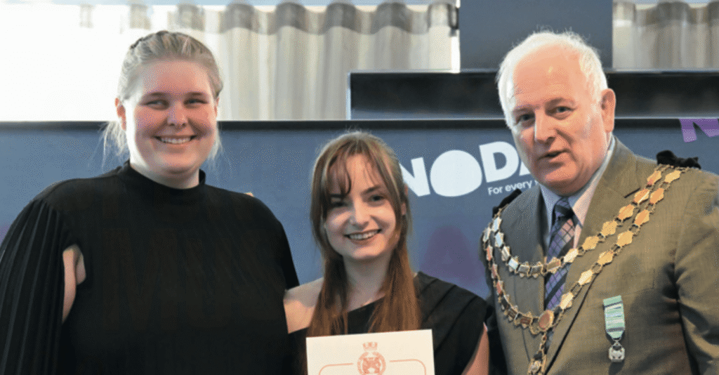 a photo showing two young women, Nadia (left) and Rowan (centre), receiving a certificate from NODA President Graham Liverton (right) in front of a blue NODA backdrop.