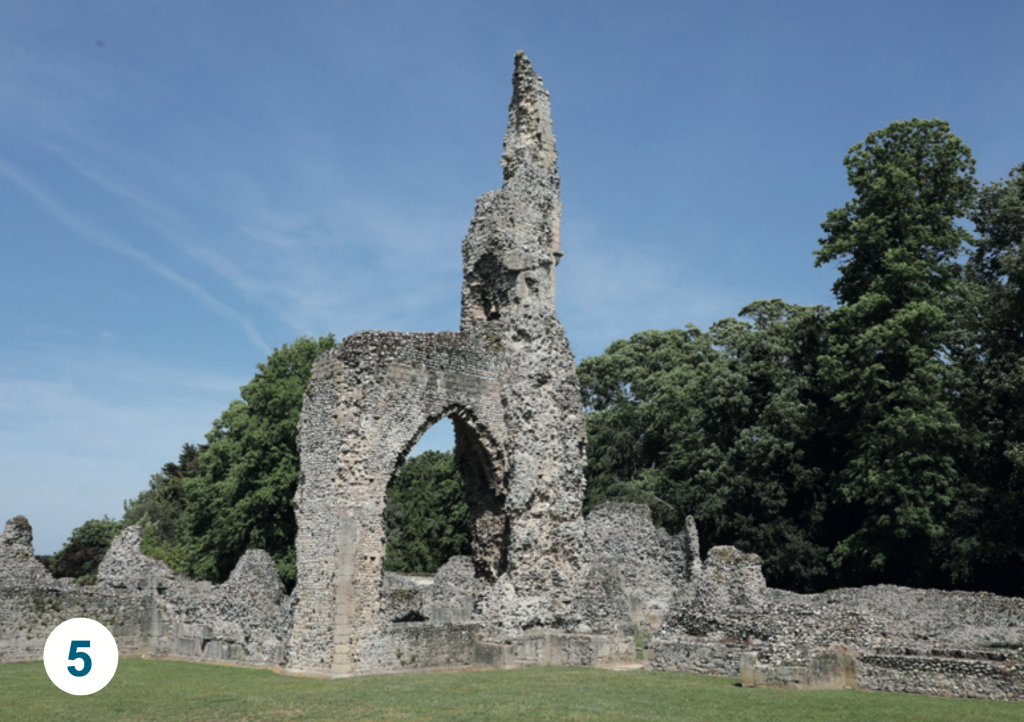 The ruins of Thetford Priory, featuring a tall surviving stone arch and medieval flint walls surrounded by grass and mature trees.