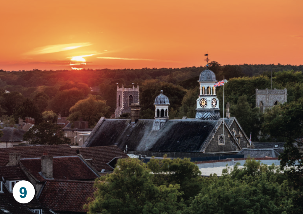 A sunset view across Thetford town centre, showing the Guildhall clock tower, church towers and rooftops silhouetted beneath a vivid orange evening sky.
