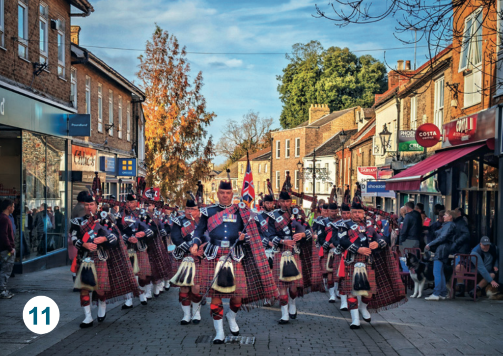 A pipe band marching through Thetford town centre, with musicians in tartan uniforms performing along the high street as spectators watch from the pavements.