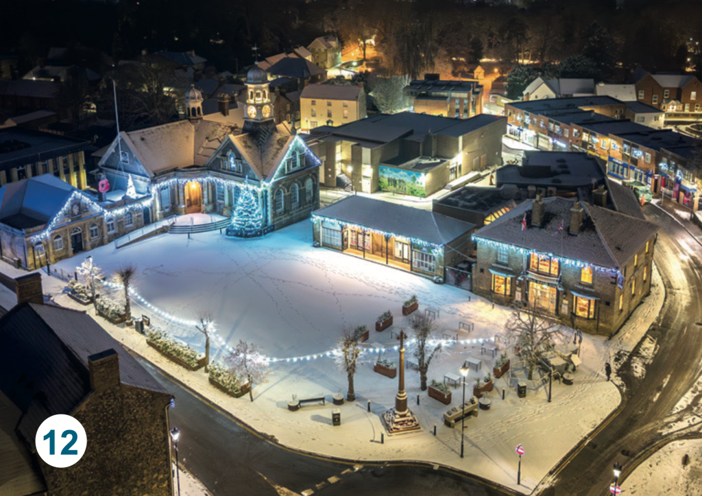 A snowy aerial view of Thetford Market Place, showing the illuminated Guildhall, Christmas lights and surrounding streets blanketed in snow at night.