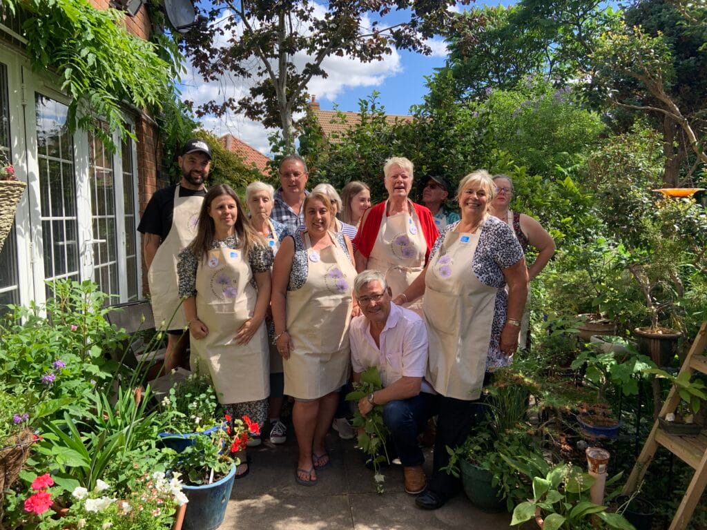 A group of volunteers wearing beige aprons with a purple flower logo pose together in a lush, green garden. The setting is bright and summery, with colourful potted plants, flowers, and hanging greenery surrounding the group. A man kneels at the front holding a plant, while others smile and stand behind him. Everyone appears ready for the Thetford Open Gardens event.