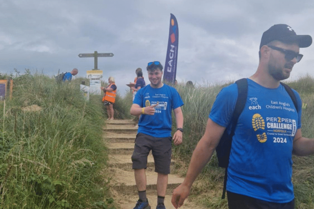 Participants in blue “Pier 2 Pier Challenge 2024” T-shirts walk down sandy steps through beach grass during the East Anglia’s Children’s Hospices (EACH) fundraiser event. One man in sunglasses is in the foreground, while another gives a thumbs up mid-step. Volunteers and signage are visible at the top of the steps beneath an overcast sky.