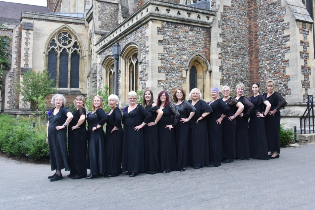 A choir of fifteen women dressed in coordinated long black outfits poses in a line outside a historic stone church with arched windows and decorative brickwork. The women, standing arm-in-arm with hands on hips, smile confidently toward the camera. Some wear sparkling tops or floral accessories. The church's intricate architecture and greenery provide a picturesque backdrop.