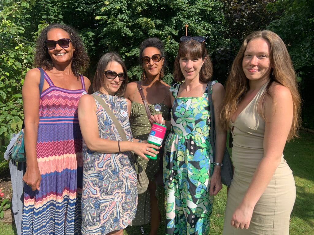 Five women stand together outdoors in a sunlit garden, smiling at the camera. They are dressed in colourful summer dresses and sunglasses, with one woman in the centre holding a green and pink St Nicholas Hospice donation pot. Behind them is lush greenery and trees.