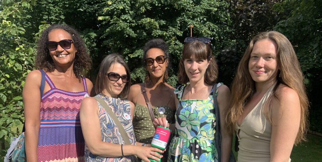 Five women stand together outdoors in a sunlit garden, smiling at the camera. They are dressed in colourful summer dresses and sunglasses, with one woman in the centre holding a green and pink St Nicholas Hospice donation pot. Behind them is lush greenery and trees.
