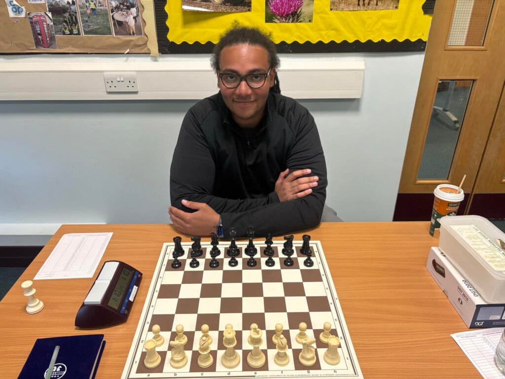 A young man with glasses and a dark jacket sits at a table with arms folded, facing a chessboard set up for play. A digital chess clock, scoresheet, and pen are on the table to his left, with a takeaway coffee cup and plastic container to his right. Behind him is a bulletin board with colourful photos and yellow backing. The setting appears to be a classroom or community room.