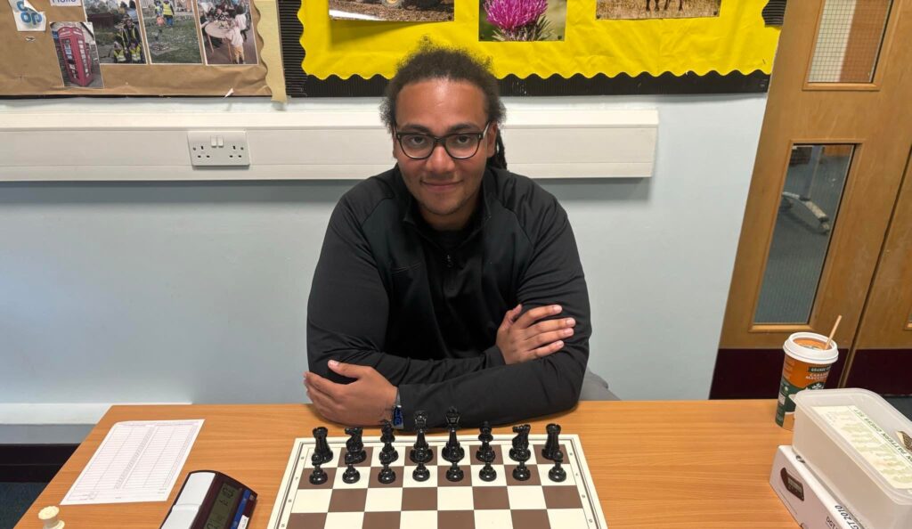 A young man with glasses and a dark jacket sits at a table with arms folded, facing a chessboard set up for play. A digital chess clock, scoresheet, and pen are on the table to his left, with a takeaway coffee cup and plastic container to his right. Behind him is a bulletin board with colourful photos and yellow backing. The setting appears to be a classroom or community room.