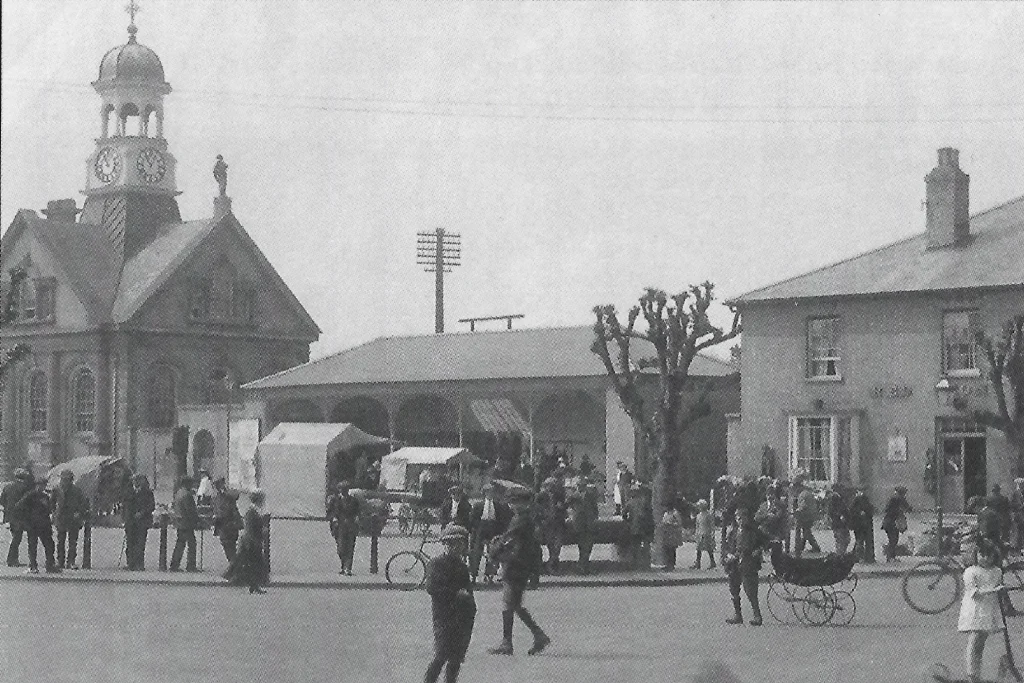 Black-and-white historic photo of Thetford Market Place, showing a busy street scene with horse-drawn prams, market stalls, and people gathered in early 20th-century dress. The Guildhall clock tower is visible on the left, with the covered market building in the centre. Trees are bare and tightly pollarded. A public house stands on the right-hand side.