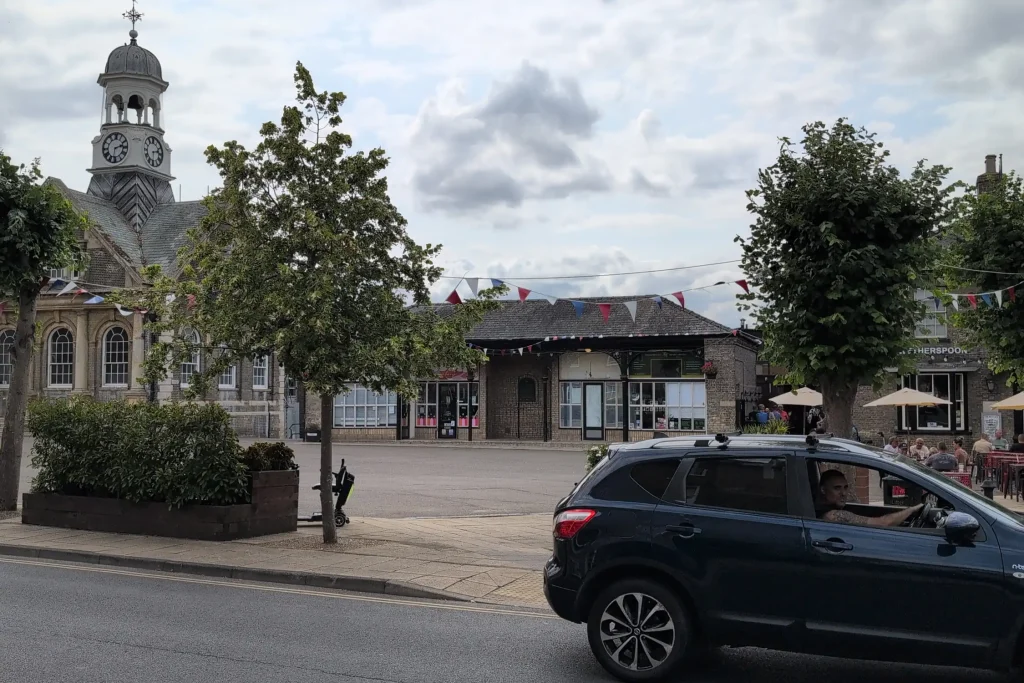 Colour photo of present-day Thetford Market Place taken from a similar angle as the historic view. The Guildhall and covered market still stand, with updated windows and bunting strung across the scene. Trees are full and leafy. A black car is passing in the foreground, and people sit outside the Wetherspoons pub under parasols. The area is clean and open with modern paving and planters.