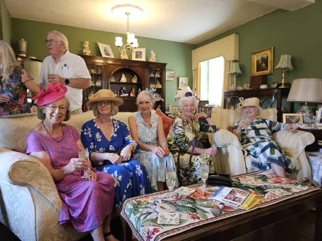 A cheerful group of women in colourful dresses and hats sit on armchairs and sofas in an elegant living room, holding glasses of bubbly. Behind them, a man in a white shirt and others mingle. The room features antique-style furniture, a green wall, framed pictures, and a display of porcelain figurines.