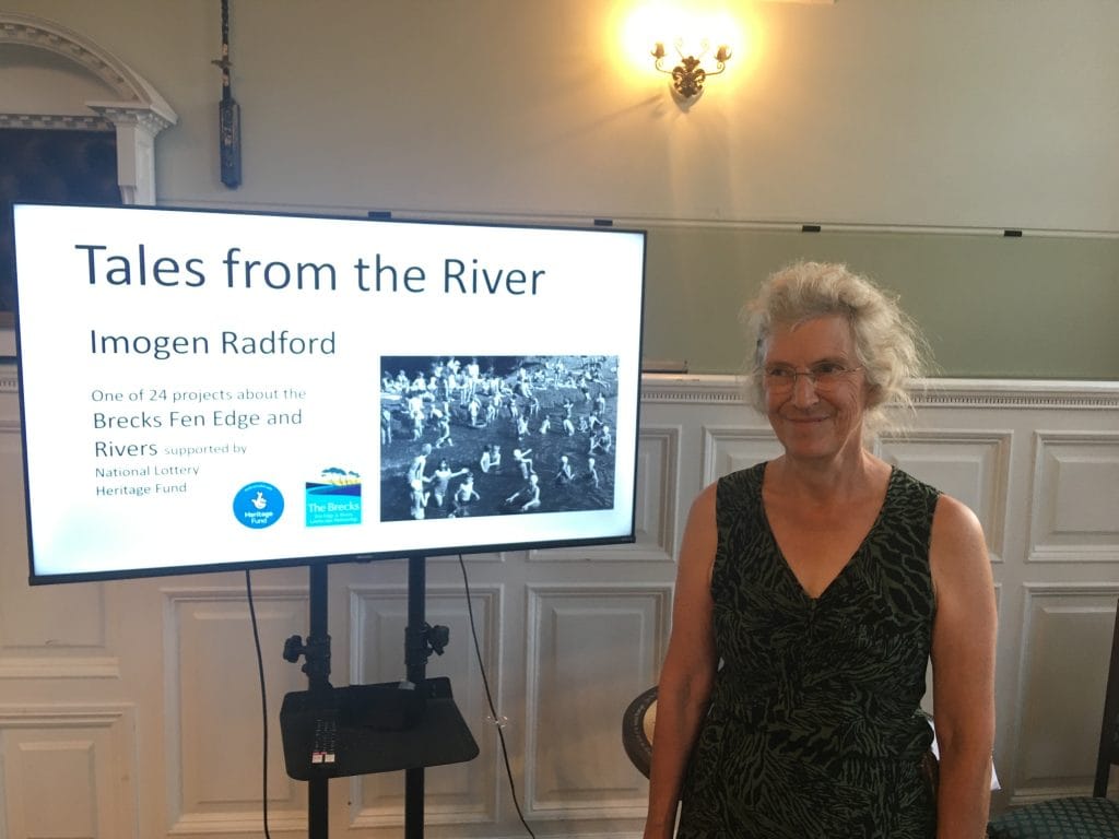 Speaker Imogen Radford stands next to a large screen displaying a presentation titled “Tales from the River.” The event is one of 24 projects supported by the Brecks Fen Edge and Rivers Landscape Partnership and the National Lottery Heritage Fund. The screen includes an old photo of people swimming or wading in a river.