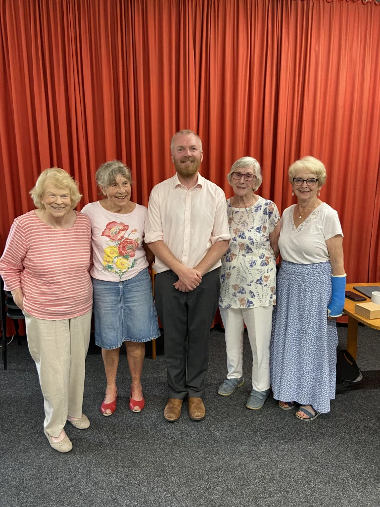 A group of five smiling people pose together indoors in front of red stage curtains. Four older women flank a younger man in a pale shirt and dark trousers. One woman has her arm in a blue cast. The setting suggests a community group or event.
