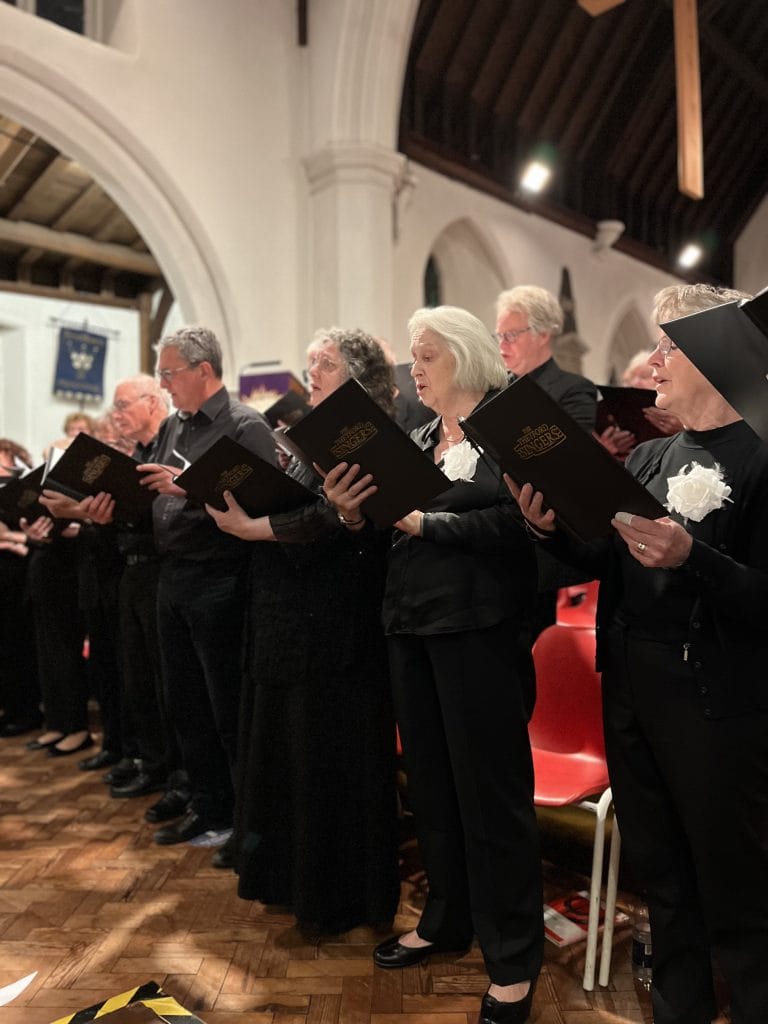 Members of the Thetford Singers choir perform inside a church with tall white arches and wooden vaulted ceilings. The choir, made up of older men and women dressed in black with white flower corsages, are holding black folders embossed with "Thetford Singers" in gold lettering. They stand in a row next to red chairs on a wooden parquet floor, singing with concentration. The atmosphere is warm and focused, with soft lighting highlighting the church's architecture and the choir’s expressions.