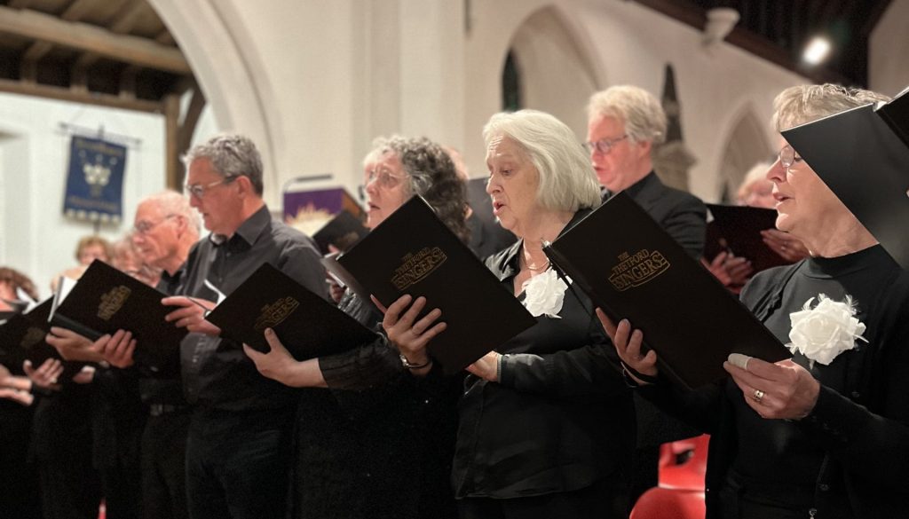 Members of the Thetford Singers choir perform inside a church with tall white arches and wooden vaulted ceilings. The choir, made up of older men and women dressed in black with white flower corsages, are holding black folders embossed with "Thetford Singers" in gold lettering. They stand in a row next to red chairs on a wooden parquet floor, singing with concentration. The atmosphere is warm and focused, with soft lighting highlighting the church's architecture and the choir’s expressions.