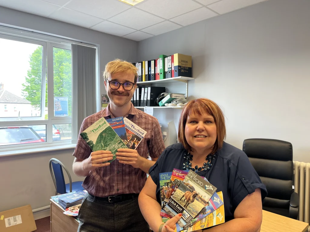 A man and woman pose in an office holding multiple past issues of About Thetford magazine. The man, wearing glasses and a checked shirt, stands by the window holding three issues. The woman,  leaning on a desk, displays a fan of colourful covers. Behind them are shelves with files and a large window letting in natural light.