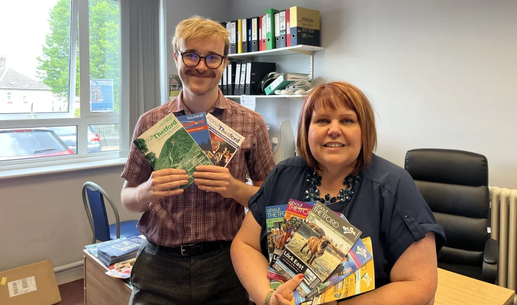 A man and woman pose in an office holding multiple past issues of About Thetford magazine. The man, wearing glasses and a checked shirt, stands by the window holding three issues. The woman, leaning on a desk, displays a fan of colourful covers. Behind them are shelves with files and a large window letting in natural light.