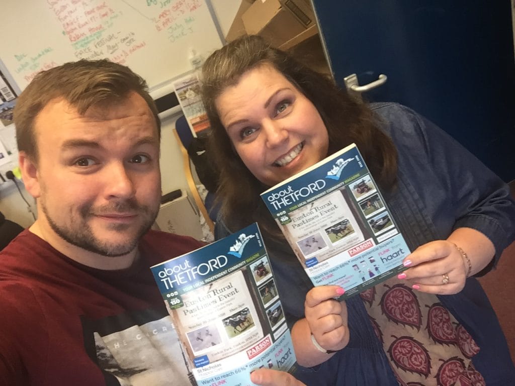 Two smiling people pose in an office holding copies of About Thetford magazine. The man wears a burgundy T-shirt, and the woman wears a navy top and patterned dress. A whiteboard with colourful writing is visible behind them, along with a desk and blue chair. The magazines feature a cover promoting the Euston Rural Pastimes Event.