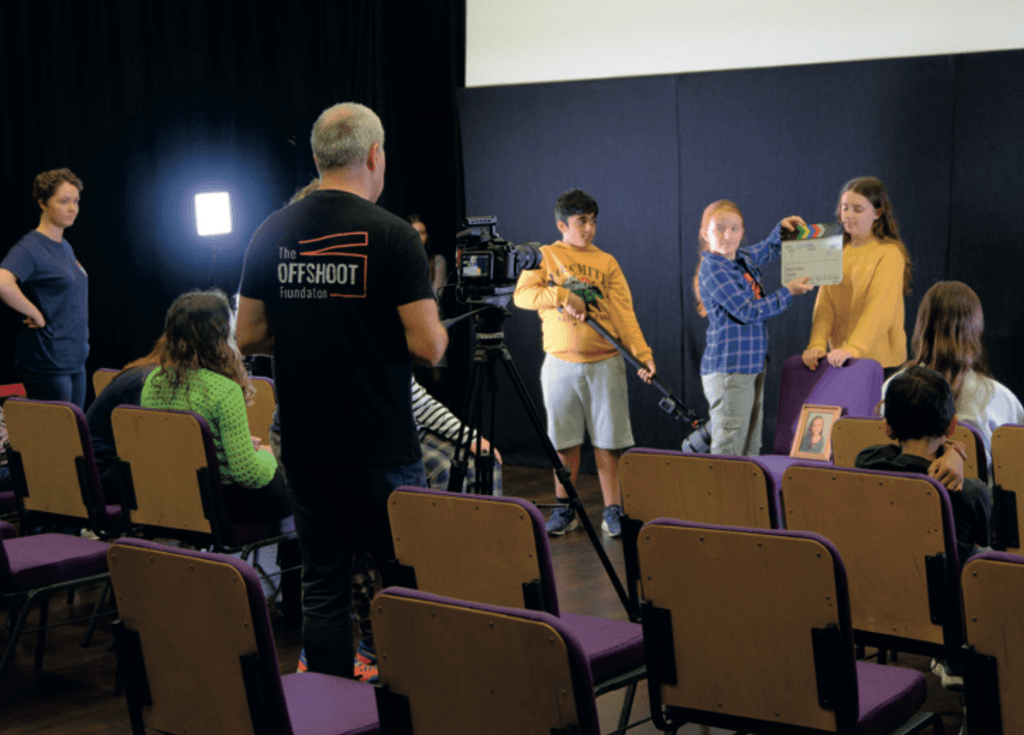A behind-the-scenes photo of young people taking part in a filmmaking workshop. A group of children are involved in a film shoot, one holding a clapperboard, another on sound boom, while others act or observe. Paul Press, founder of The Offshoot Foundation, stands by the camera wearing a black “OFFSHOOT Foundation” t-shirt. The session takes place in a darkened room with theatre-style chairs and professional lighting equipment.