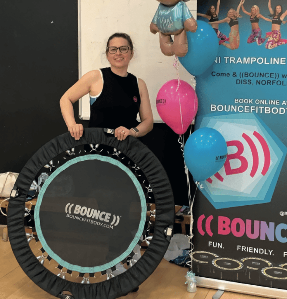 Bounce instructor Giorgiana stands smiling, holding a mini trampoline branded with the “((BOUNCE))” logo. She’s dressed in sportswear and ready to teach a fitness class. Bright balloons and a colourful promotional banner are in the background, featuring the BOUNCEFITBODY.com website and more class branding.