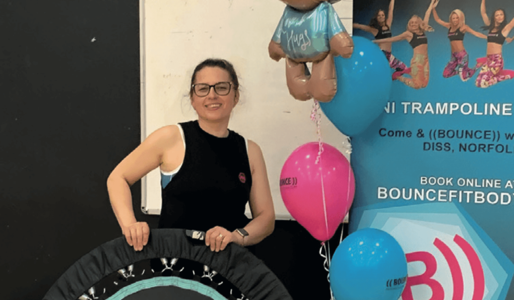 Bounce instructor Giorgiana stands smiling, holding a mini trampoline branded with the “((BOUNCE))” logo. She’s dressed in sportswear and ready to teach a fitness class. Bright balloons and a colourful promotional banner are in the background, featuring the BOUNCEFITBODY.com website and more class branding.