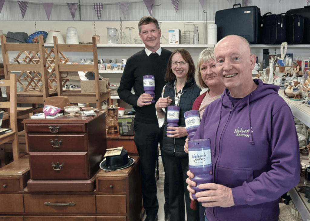 Four people standing inside a Norfolk Reuse Shop, smiling and holding purple charity collection tins for Nelson’s Journey. The man in front wears a branded purple hoodie with the Nelson’s Journey logo. Behind them, shelves and tables are filled with second-hand items including furniture, crockery, ornaments, bags, and toys. Bunting is hung above the shelves, adding to the welcoming, community-focused atmosphere.