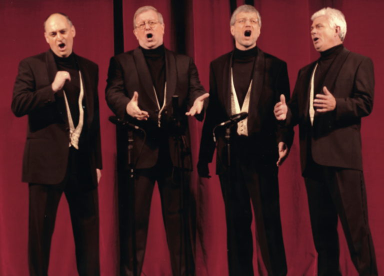 Four men wearing matching black suits and light-coloured waistcoats perform in front of a red curtain. Each sings passionately into microphones, part of a barbershop-style vocal group.