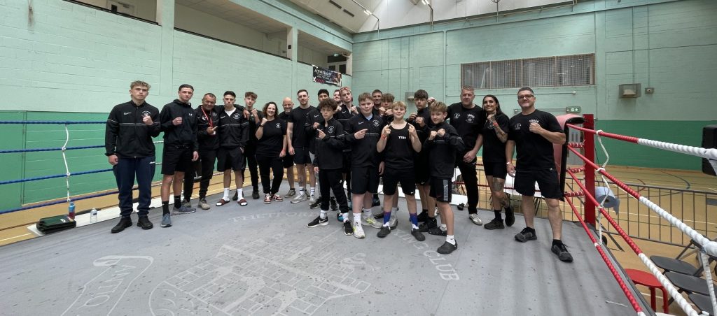 Group photo of approximately 22 members of a boxing club—mostly teenage boys along with a few adult coaches—posing together in a boxing ring inside a large indoor sports hall. Everyone is dressed in black sportswear branded with the club logo. Several of the boxers are holding up their fists in a classic fighter’s pose. The ring floor features a faint crest and the words “THETFORD BOXING CLUB.” The hall has high ceilings with skylights and green-and-white walls, giving a bright and spacious atmosphere.