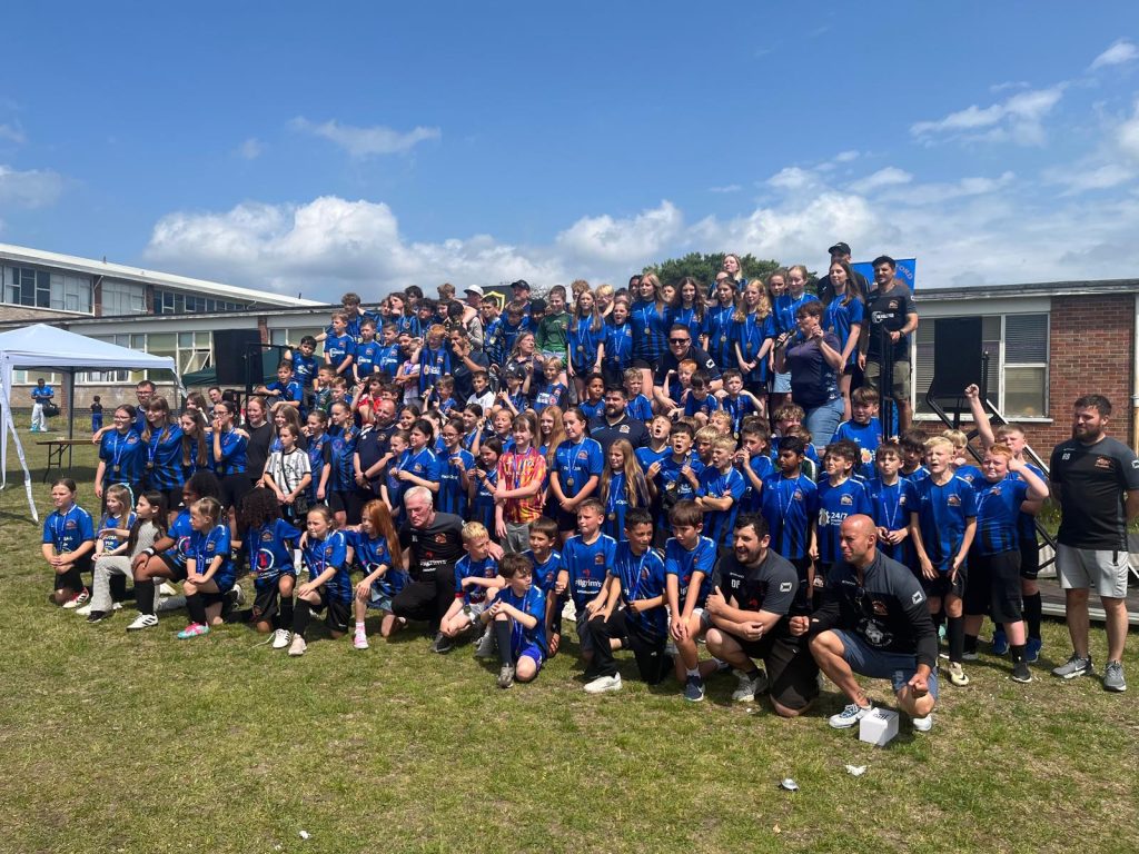 Large group photo of Thetford Bulldogs youth footballers gathered outside on a sunny day. Dozens of children in matching blue team shirts stand and kneel in rows, with coaches and adults alongside them. They are smiling, cheering, and posing for the picture on the grass, with a school building and gazebo in the background.