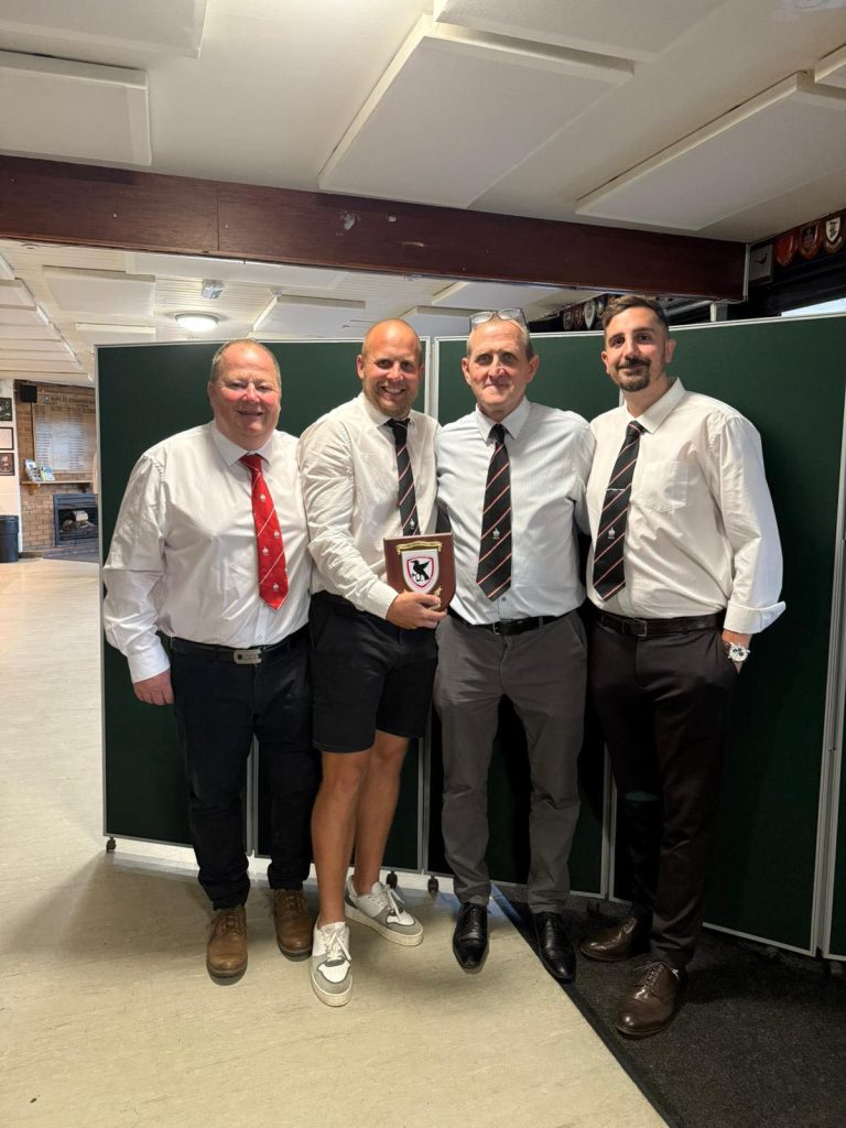 Four men in white shirts and black-and-red striped ties pose indoors, one holding a wooden plaque award.