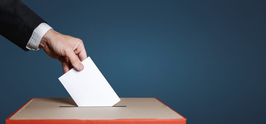 A hand in a dark suit jacket places a white ballot paper into a cardboard ballot box with a slot. The background is a solid dark blue.